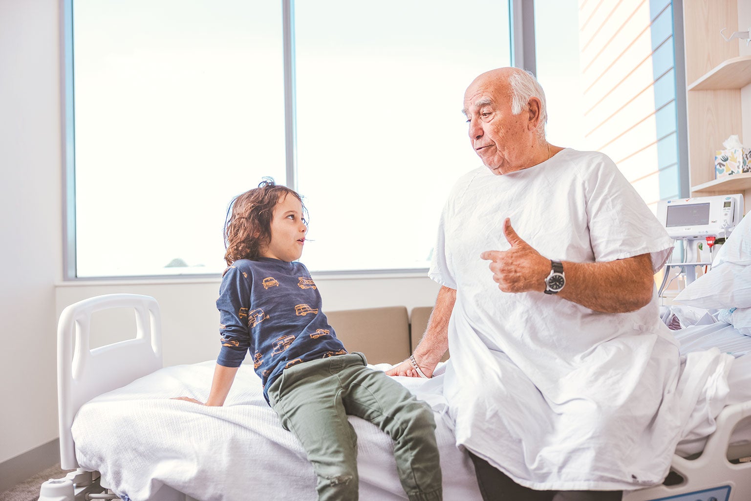 older man in hospital gown talking to child on hospital bed